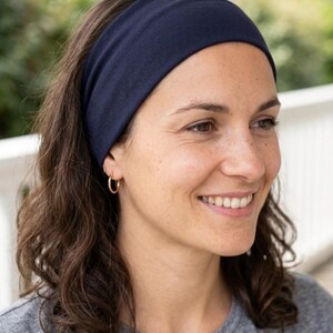 Peut inclure: Un large bandeau en tissu bleu marine uni, fabriqué dans une matière douce et extensible. Le bandeau est porté sur le front et repose fermement autour de la tête, présentant une finition lisse et mate ainsi qu'un design simple et épuré.