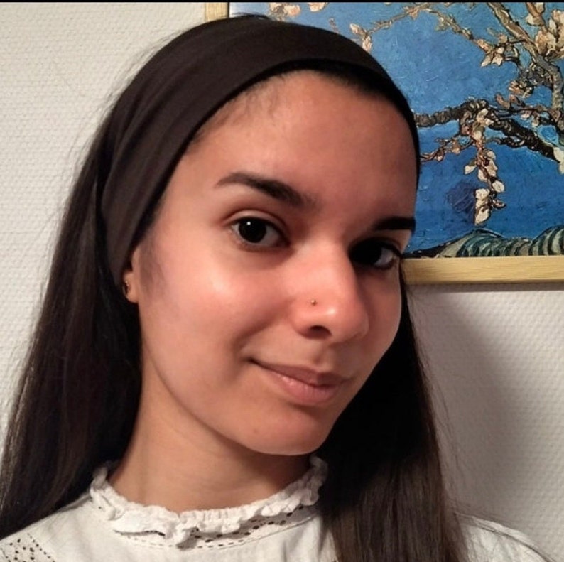 May include: A woman with long dark hair is wearing a brown headband and a white blouse with a lace collar. She has a nose ring and is looking at the camera.