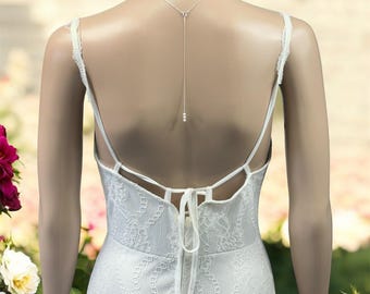 May include: A white lace dress with a low back, thin straps, and a tie closure at the waist. A delicate silver-toned chain necklace hangs down the center of the back, featuring a small pendant with three tiny white beads at the bottom end.