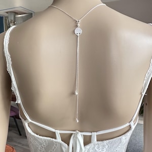 Może przedstawiać: Srebrny naszyjnik z tyłu z zawieszką w kształcie drzewa życia i dwoma akcentami pereł. Naszyjnik znajduje się na białej koronkowej sukience.