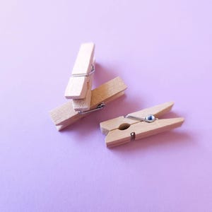 Könnte beinhalten: Zwei Holzklammern auf einem hellvioletten Hintergrund. Eine Klammer liegt auf der anderen, und die dritte Klammer liegt flach.