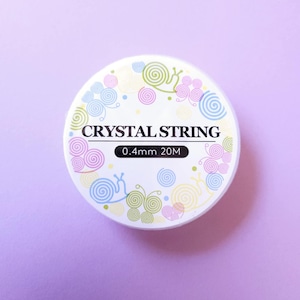 May include: A white spool of crystal string with the text "CRYSTAL STRING" in black. The spool is decorated with colorful snail and swirl designs. The label reads "0.4mm 20M". The background is a solid light purple.