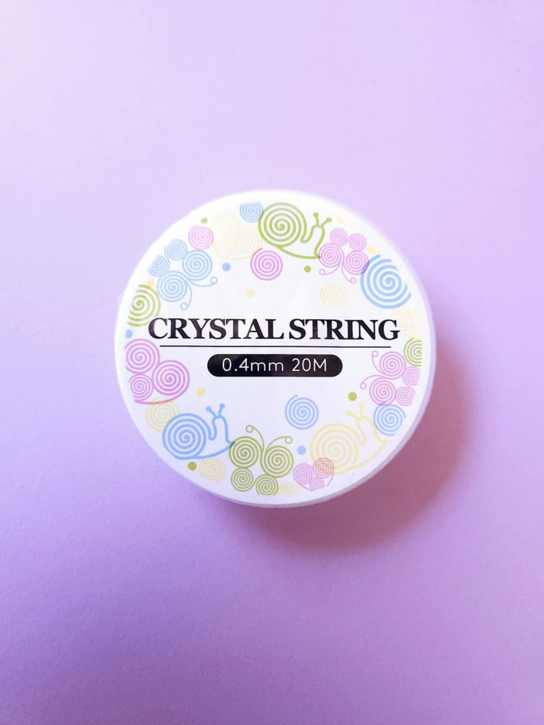 Puede incluir: Una bobina blanca de hilo de cristal con el texto "CRYSTAL STRING" en negro. La bobina est&aacute; decorada con coloridos dise&ntilde;os de caracoles y espirales. La etiqueta dice "0,4 mm 20 M". El fondo es de color lila claro s&oacute;lido.