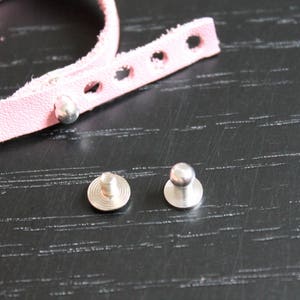 Può includere: Due bottoni a pressione in metallo argentato, uno con la parte superiore piatta e l'altro con la parte superiore arrotondata, su una superficie nera.