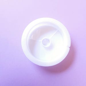 Puede incluir: Un objeto de pl&aacute;stico blanco con forma circular y un peque&ntilde;o agujero en el centro. El objeto tiene un borde elevado y una superficie lisa y limpia. El fondo es de color lila claro.