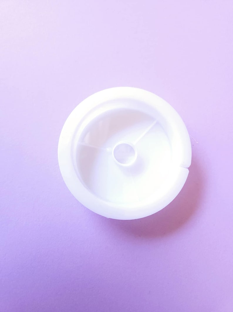 Puede incluir: Un objeto de pl&aacute;stico blanco con forma circular y un peque&ntilde;o agujero en el centro. El objeto tiene un borde elevado y una superficie lisa y limpia. El fondo es de color lila claro.