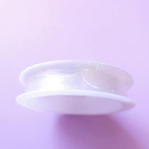 Puede incluir: Un carrete de hilo transparente y fino para abalorios, enrollado firmemente en una bobina de pl&aacute;stico blanco. La bobina tiene una base circular plana y un borde ligeramente elevado. El fondo es de color p&uacute;rpura claro.
