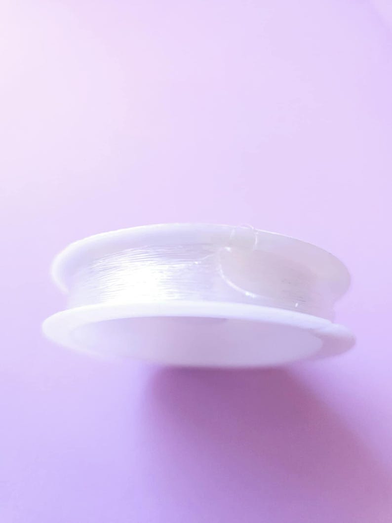 Puede incluir: Un carrete de hilo transparente y fino para abalorios, enrollado firmemente en una bobina de pl&aacute;stico blanco. La bobina tiene una base circular plana y un borde ligeramente elevado. El fondo es de color p&uacute;rpura claro.