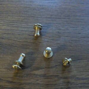 May include: Four gold-toned metal fasteners with different designs. Two have a round head and a small metal ring, one has a round head and a screw, and one has a screw and a flat head.