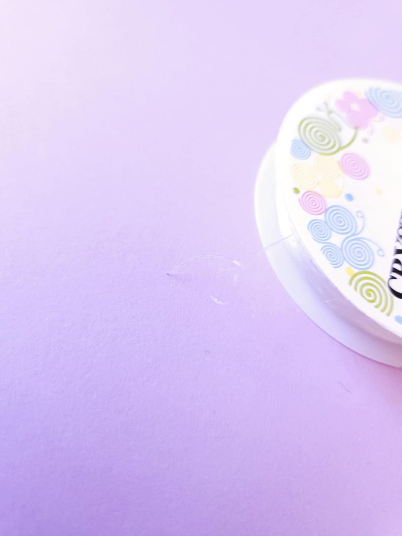 Puede incluir: Un carrete de hilo de abalorios transparente y fino es parcialmente visible sobre un fondo morado claro. El borde de un carrete blanco con un dise&ntilde;o floral y en espiral est&aacute; en la esquina superior derecha. El hilo se utiliza para hacer joyas.