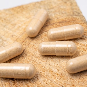 May include: Six beige capsules on a light brown wooden surface. The capsules are translucent and show a light brown powder inside.