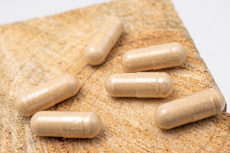 May include: Six beige capsules on a light brown wooden surface. The capsules are translucent and show a light brown powder inside.