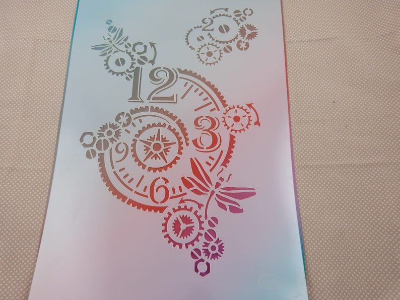 May include: A white stencil with a clock face design featuring gears, a dragonfly, and the numbers 12, 3, 6, and 9.