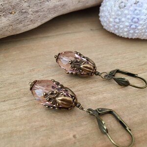 Pode incluir: Um par de brincos pendentes com uma conta de vidro ovalada rosa claro e detalhes em metal de tom dourado antigo. Os brincos t&ecirc;m um fecho de gancho.
