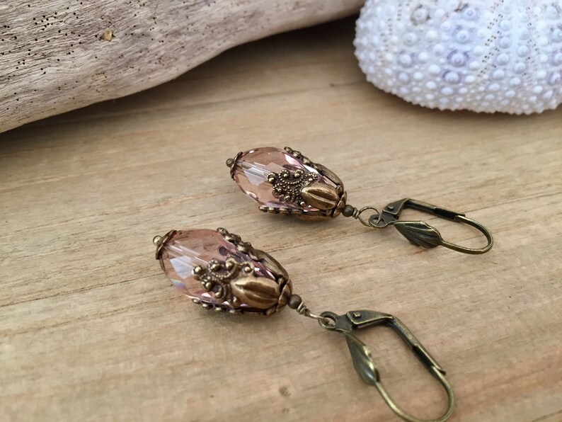 Pode incluir: Um par de brincos pendentes com uma conta de vidro ovalada rosa claro e detalhes em metal de tom dourado antigo. Os brincos t&ecirc;m um fecho de gancho.