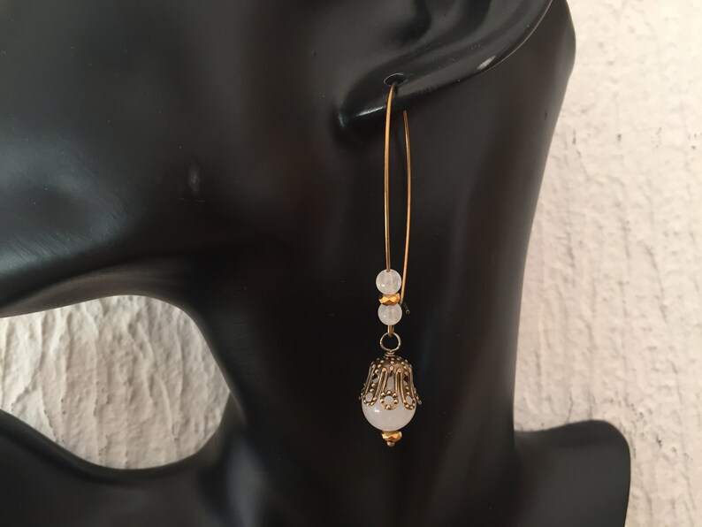Pode incluir: Brincos de argola dourados com uma pedra branca e detalhes de contas douradas.