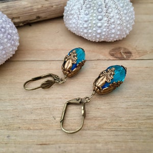 Pode incluir: Um par de brincos com contas de vidro azuis e detalhes em metal dourado. As contas s&atilde;o de forma oval e t&ecirc;m um design facetado. Os brincos est&atilde;o pendurados em ganchos dourados.