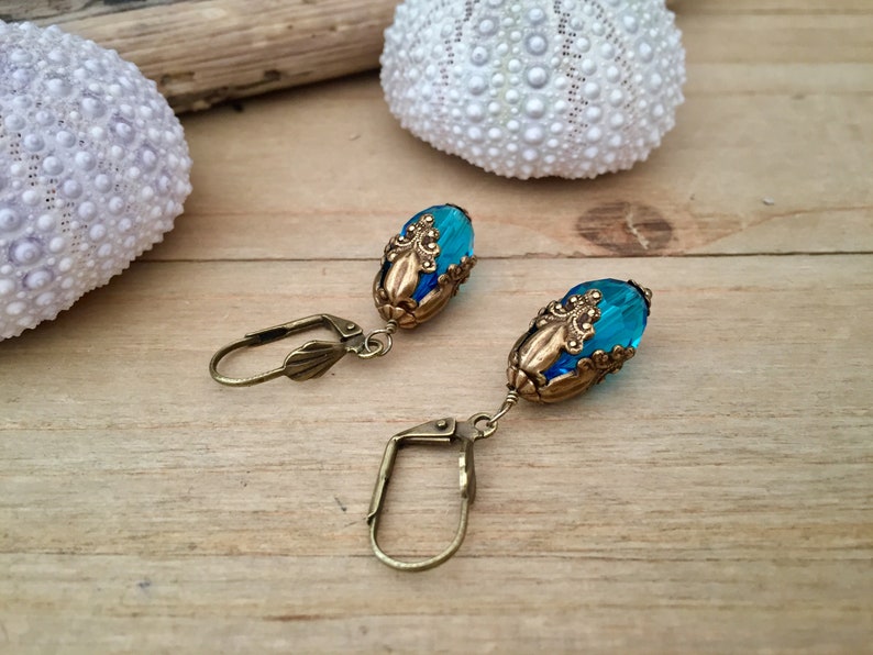 Pode incluir: Um par de brincos com contas de vidro azuis e detalhes em metal dourado. As contas s&atilde;o de forma oval e t&ecirc;m um design facetado. Os brincos est&atilde;o pendurados em ganchos dourados.