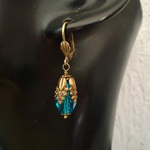 Pode incluir: Um par de brincos pendentes com uma conta de cristal azul turquesa e uma moldura de metal dourado adornada.