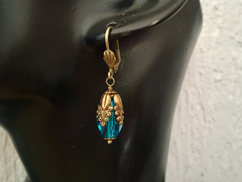 Pode incluir: Um par de brincos pendentes com uma conta de cristal azul turquesa e uma moldura de metal dourado adornada.