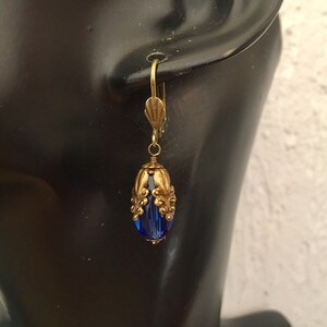 Pode incluir: Um par de brincos dourados com uma conta de vidro azul em forma de l&aacute;grima. A conta &eacute; cercada por uma filigrana decorativa dourada.