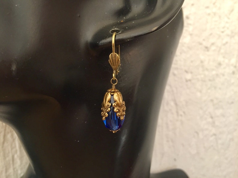 Pode incluir: Um par de brincos dourados com uma conta de vidro azul em forma de l&aacute;grima. A conta &eacute; cercada por uma filigrana decorativa dourada.