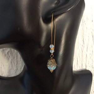 Pode incluir: Um par de brincos de argola dourados com uma conta de vidro azul claro e um design de filigrana dourada. Os brincos est&atilde;o pendurados na orelha de um manequim.