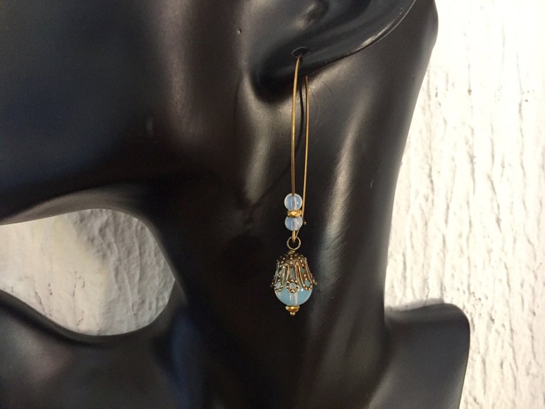 Pode incluir: Um par de brincos de argola dourados com uma conta de vidro azul claro e um design de filigrana dourada. Os brincos est&atilde;o pendurados na orelha de um manequim.