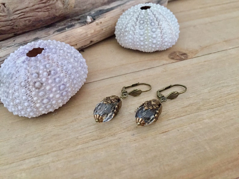 Pode incluir: Um par de brincos de bronze com contas de vidro transparente em forma de l&aacute;grima. As contas t&ecirc;m uma superf&iacute;cie texturizada e s&atilde;o adornadas com desenhos florais de bronze.