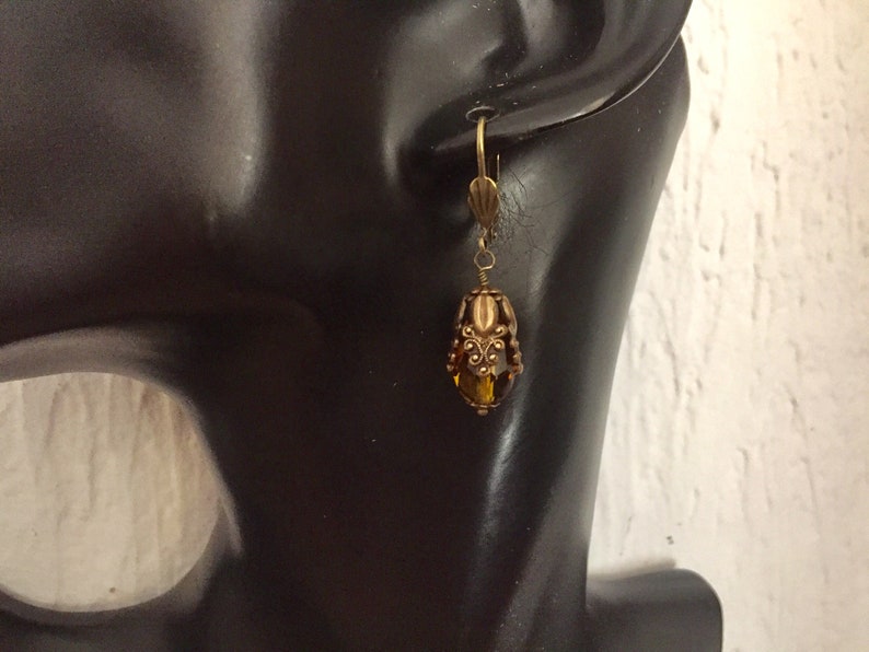 Pode incluir: Um par de brincos de argola dourados com uma conta de vidro marrom no centro. A conta &eacute; cercada por uma moldura de metal decorativa.