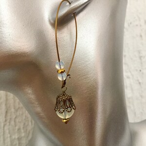 Pode incluir: Brincos de argola dourados com uma conta de vidro branca e um pingente de filigrana dourada.
