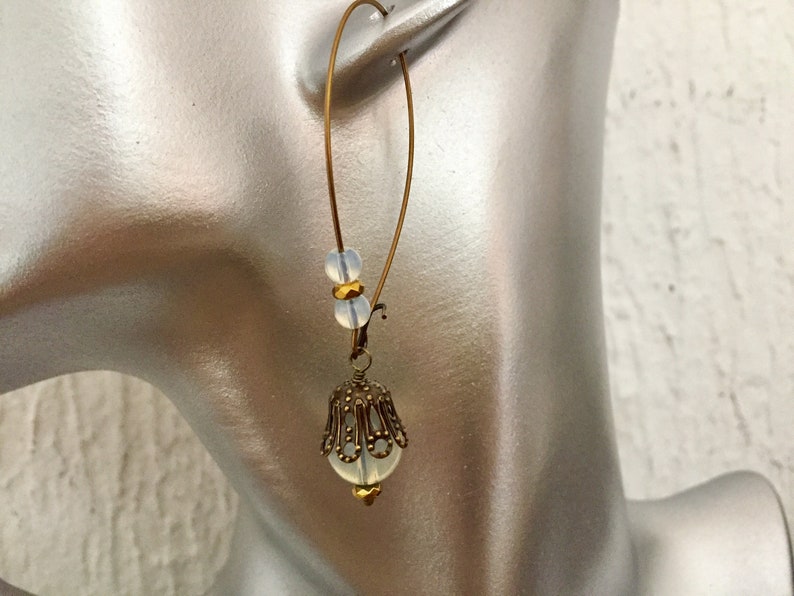 Pode incluir: Brincos de argola dourados com uma conta de vidro branca e um pingente de filigrana dourada.