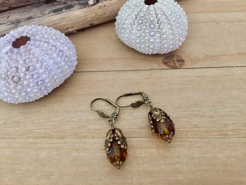 Pode incluir: Um par de brincos de bronze com contas de vidro &acirc;mbar em forma de l&aacute;grima. Os brincos t&ecirc;m um design floral detalhado.