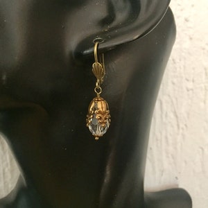 Pode incluir: Brincos de argola de tom dourado com uma conta de cristal transparente e detalhes ornamentados.