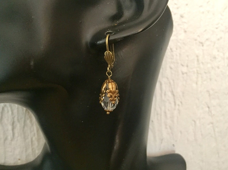 Pode incluir: Brincos de argola de tom dourado com uma conta de cristal transparente e detalhes ornamentados.