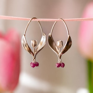 Pode incluir: Um par de brincos de argola prateados com um elemento central de metal em forma de tulipa e duas pequenas contas de pedra preciosa facetadas em rosa escuro penduradas na base, suspensos por uma fita fina de cor rosa claro.