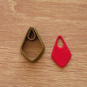 May include: A brown metal earring-shaped cookie cutter next to a red earring. The cutter is a teardrop shape with a smaller teardrop cutout at the top. The red earring is a solid teardrop shape with a loop at the top.