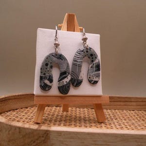 Peut inclure: Une paire de boucles d'oreilles pendantes en forme d'arche avec un motif monochrome de tourbillons, de points et de lignes noirs, blancs et gris. Les boucles d'oreilles sont suspendues à des crochets en métal argenté sur une toile blanche.