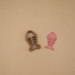 May include: A bronze-colored fish skeleton cookie cutter with a textured surface and a separate pink fish skeleton cutout shape. The cookie cutter features a hollow center with rib-like details, while the pink cutout is a solid, flat piece.