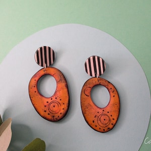 Puede incluir: Un par de pendientes de declaración naranja y negro con un diseño geométrico. Los pendientes presentan una forma ovalada grande con un círculo más pequeño cortado en el centro. La parte superior de cada pendiente es un círculo de rayas blancas y negras.