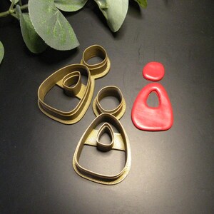 Puede incluir: Dos cortadores de pendientes de plástico de color dorado con un marco exterior en forma de lágrima y una sección superior circular, acompañados de una pieza de arcilla roja que consiste en un círculo pequeño y una lágrima más grande.