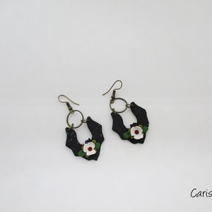 Op de afbeelding: Een paar zwarte vleermuisvormige oorbellen met witte bloemen en groene bladeren. De oorbellen hebben een gouden ring aan de bovenkant.