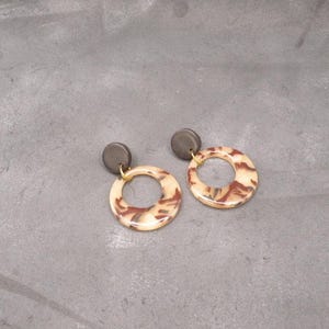 Puede incluir: Un par de pendientes con diseño circular y un patrón veteado en crema, marrón y beige. Los pendientes tienen un perno gris oscuro y un lazo de conexión dorado. Los pendientes se muestran sobre una superficie gris.