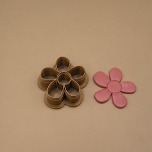 Peut inclure: Un emporte-pièce en forme de fleur de couleur bronze avec un centre creux et cinq pétales arrondis, placé à côté d'une découpe en forme de fleur rose faite d'un matériau lisse et plat qui correspond à la forme du design intérieur de l'outil.