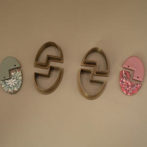 May include: Two oval-shaped clay earring components and two matching oval-shaped plastic cutters. One earring set is solid sage green and patterned white and green, while the other is solid dusty pink and patterned pink, white, and dark red.