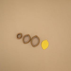 May include: Three brown, lemon-shaped cookie cutters of varying sizes arranged in a row, accompanied by a single, solid yellow, lemon-shaped cutout piece made of a smooth material, all displayed against a plain, neutral-toned background.