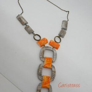 Può includere: Una collana argentata e arancione con una catena e una serie di perline quadrate. Le perline sono collegate da una catena argentata. La collana è su uno sfondo bianco.
