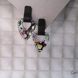 Peut inclure: Une paire de boucles d'oreilles en forme de cœur noires et blanches avec un motif abstrait coloré. Les boucles d'oreilles ont une boucle en métal noir en haut.