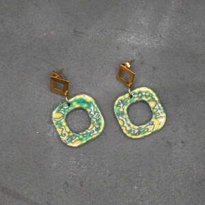 Peut inclure: Une paire de boucles d'oreilles avec un dessus carré doré et un pendentif circulaire vert et jaune. Le pendentif a une forme organique unique avec un trou au centre. Les boucles d'oreilles sont posées sur un fond gris.