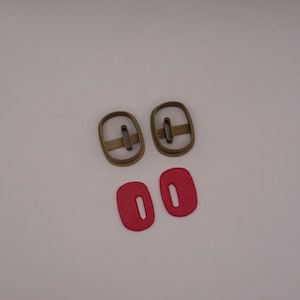 Può includere: Due set di forniture per l'artigianato sono esposti su sfondo bianco. Due stampi per fibbie ovali in metallo color bronzo sono posizionati sopra due pezzi di pelle ovali rossi con una fessura centrale. Questi articoli sono probabilmente per la lavorazione della pelle o progetti simili.
